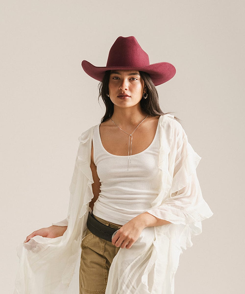 Woman wearing a white top, beige pants, and a red cowboy hat against a plain background